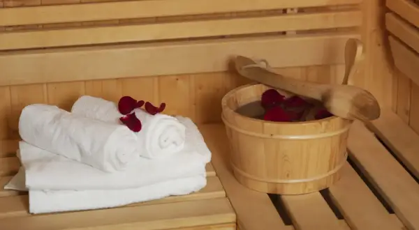 Białe ręczniki i drewniane wiaderko z wodą i płatkami róż w saunie. Dylemat: najpierw sauna sucha czy parowa?