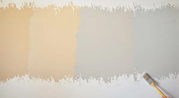 Échantillons de peinture sur un mur texturé, montrant 2 ou 3 couches de peinture dans des tons de beige et de gris. Un pinceau est visible à droite.