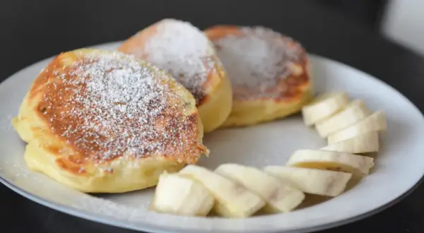 Szybkie placuszki na śniadanie, posypane cukrem pudrem, podane z plasterkami banana na białym talerzu.