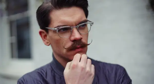 Un homme au visage losange, lunettes fines et moustache élégante, réfléchit.