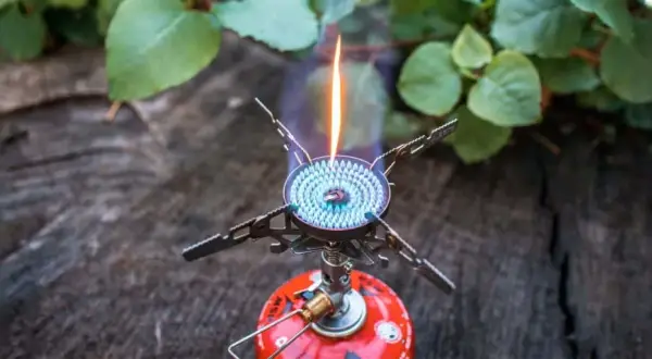 Soto Windmaster Kocher mit blauer Flamme auf einem Baumstamm, umgeben von grünen Blättern.