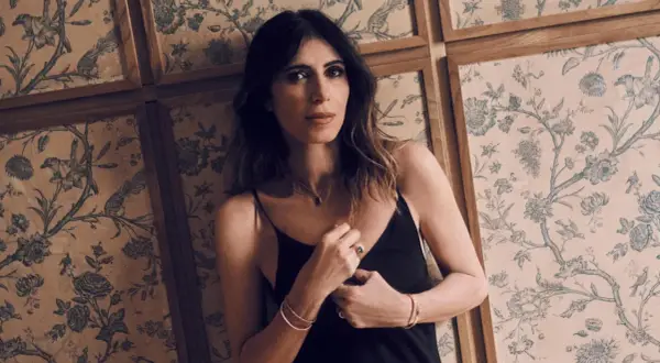 Woman in black slip dress leans against a wall with floral wallpaper.