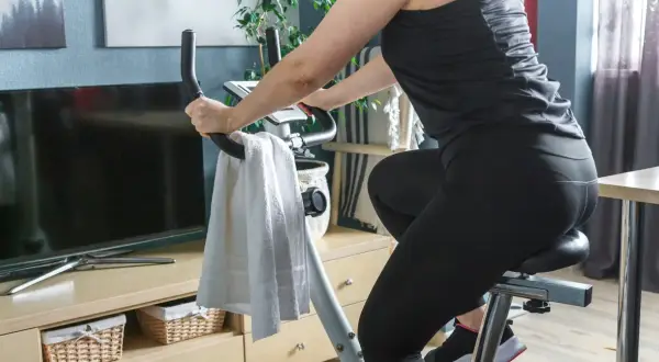 Kobieta ćwiczy na rowerze stacjonarnym. Widać jej nogi w czarnych legginsach i ręce na kierownicy. Obok wisi biały ręcznik. Jazda na rowerze stacjonarnym efekty po miesiącu są widoczne!