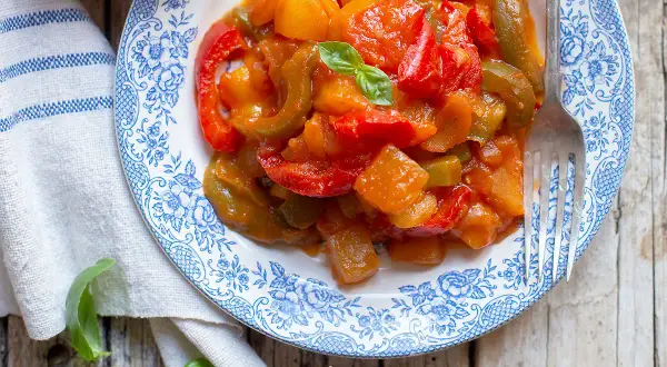Piatto di peperonata con patate, colorata e saporita, guarnita con basilico fresco su un piatto decorato.