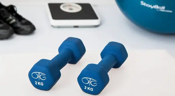 Dwa niebieskie hantle po 2 kg, waga, buty sportowe i piłka gimnastyczna.