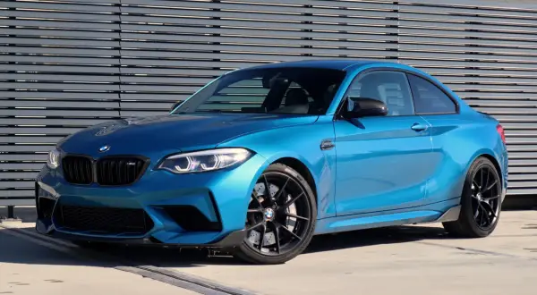 Błękitne BMW M2 Competition z czarnymi felgami i detalami, zaparkowane przed nowoczesną, szarą ścianą.