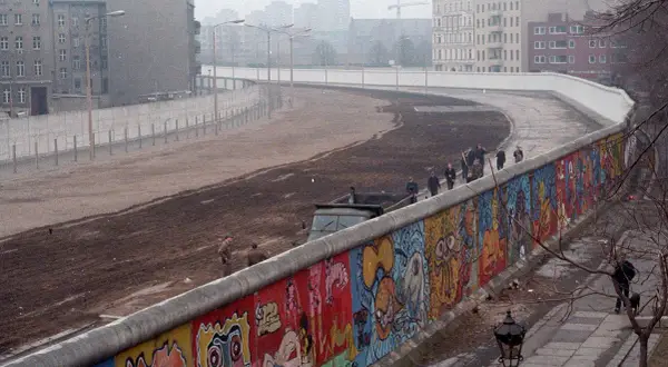 Kolorowy Mur Berliński, świadectwo czasów, kiedy powstał mur berliński, z ludźmi spacerującymi po jego szczycie i budynkami w tle.
