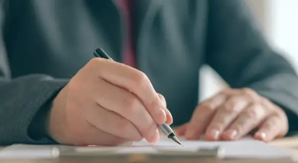 Dłoń w garniturze pisze piórem na dokumencie, co symbolizuje postępowanie egzekucyjne w administracji.