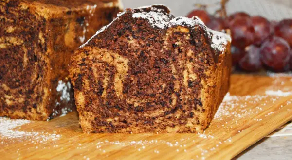 Proste ciasto bananowe z olejem i kakao, z widocznym marmurkowym wzorem. Posypane cukrem pudrem, obok kiść winogron.