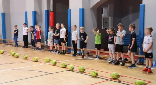 Dzieci stoją w rzędzie na sali gimnastycznej, gotowe do prostych konkurencji sportowych. Przed nimi leżą żółte piłki.