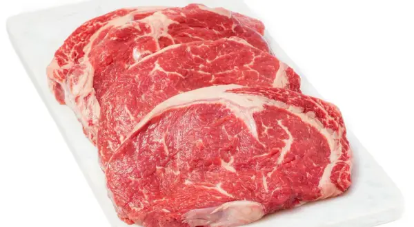Tre råa, marmorerade entrecôte-biffar på en vit skärbräda, redo för medium rare temp.