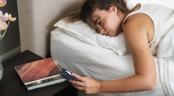 Kobieta leży w łóżku, patrząc na telefon. Obok książka i kwiaty. Czy to odzwierciedlenie współczesnego tempa życia?