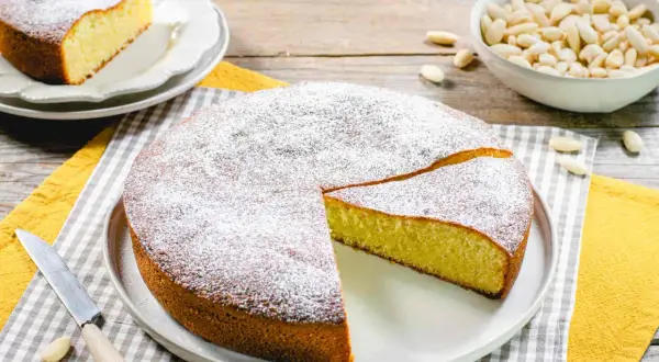 Torta soffice con farina di mandorle, spolverata di zucchero a velo. Accanto, una ciotola di mandorle intere e una fetta già servita.