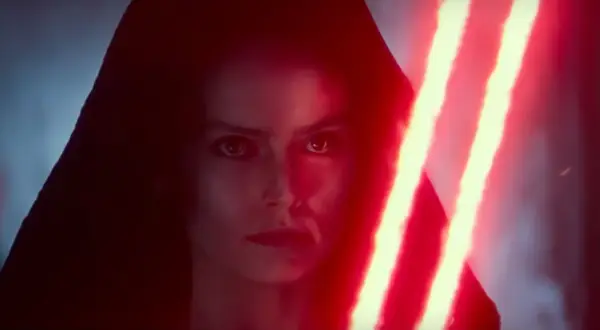 Rey Skywalker w kapturze, z ponurym wyrazem twarzy, trzyma podwójny czerwony miecz świetlny.