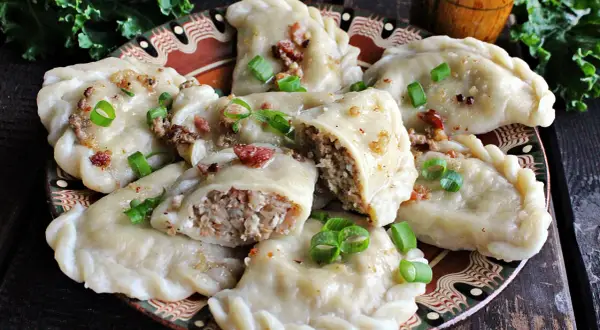 Pyszne pierogi z kaszą gryczaną, okraszone skwarkami i szczypiorkiem, na ozdobnym talerzu.