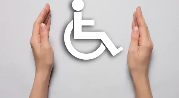 Dwie dłonie otaczają biały symbol osoby na wózku inwalidzkim na jasnoszarym tle.