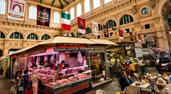 Interno di un vivace mercato delle vettovaglie, con banchi di carne, prodotti freschi e persone che fanno acquisti.