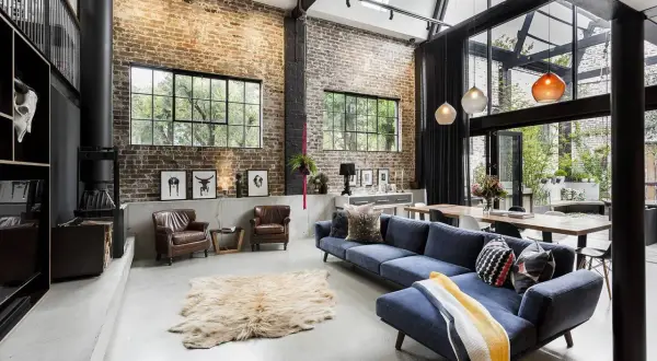 A spacious living area with exposed brick walls, concrete floors, and industrial interior design elements. A blue sectional sofa, leather armchairs, and a large dining table create a cozy yet modern atmosphere.