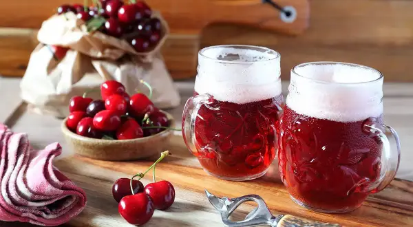 Due boccali di birra kriek rossastra con schiuma bianca, circondati da ciliegie fresche su un tagliere di legno.