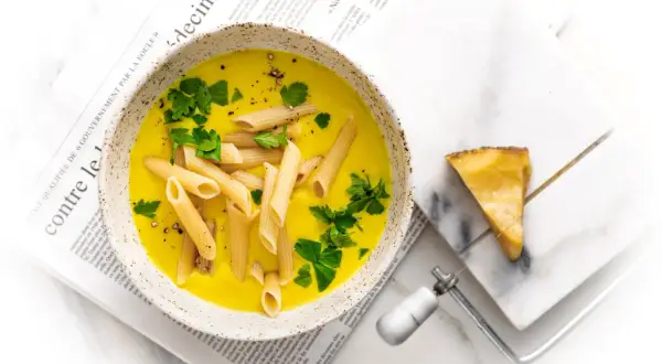 Zupa neapolitańska z makaronem penne i natką pietruszki, podana na gazecie. Obok kawałek sera i serownik.