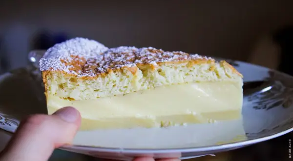 Une part de gâteau magique vanille, saupoudrée de sucre glace, repose sur une assiette. La crème onctueuse contraste avec la texture aérienne du biscuit.