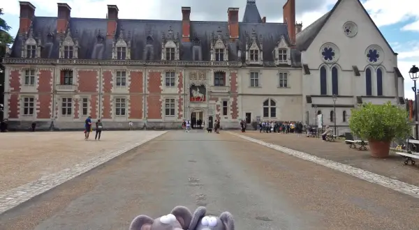 Deux souris en peluche devant le majestueux **château de Blois**. Des visiteurs explorent la cour de ce monument historique.