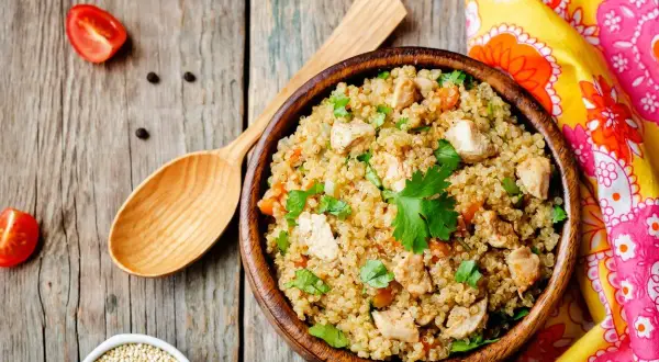 Una ciotola di quinoa con pollo e verdure, un piatto ricco di quinoa valori nutrizionali, su un tavolo di legno con pomodorini e un cucchiaio.