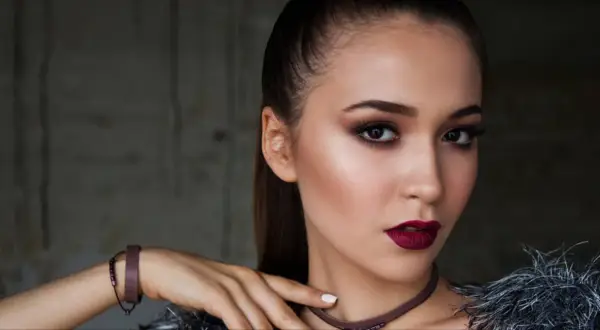 Kobieta z wyrazistym makijażem wieczorowym, ciemnymi ustami i smoky eyes. Długie włosy spięte w kucyk.