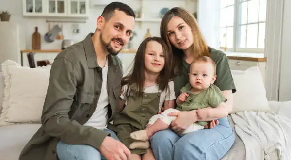 Szczęśliwa rodzina z dwójką dzieci siedzi na kanapie. Rodzice i dzieci uśmiechają się. Program 800 plus obejmuje dzieci do 18 roku życia.