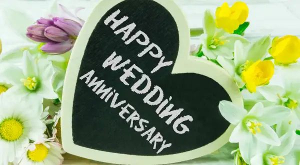 Cuore nero con scritta "Happy Wedding Anniversary" circondato da fiori bianchi, gialli e viola. Un'idea romantica per sapere dove festeggiare anniversario matrimonio.