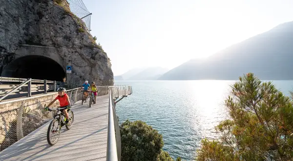 Ciclisti su una pista ciclabile panoramica lungo il lungolago di Desenzano, con un tunnel scavato nella roccia e il lago scintillante.