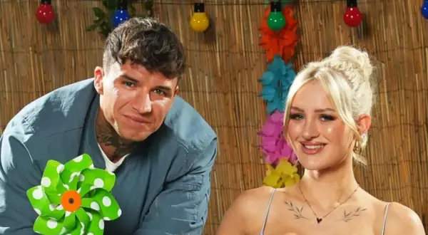 Ein Mann mit Tätowierungen und eine Frau mit blonden Haaren lächeln vor einer Bambuswand mit bunten Lichtern und Blumen. Sie sind bereit für das Sommerhaus der Stars 2025.