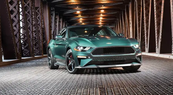 En grön pony car, en Ford Mustang, står på ett gallergolv under en bro med belysning.