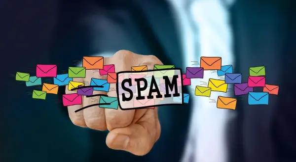 Dłoń wskazuje na napis SPAM otoczony kolorowymi kopertami, symbolizującymi niechciane wiadomości. To definicja spamu.