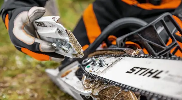 Persona con guantes realiza mantenimiento de motosierra, ajustando piezas cubiertas de serrín.