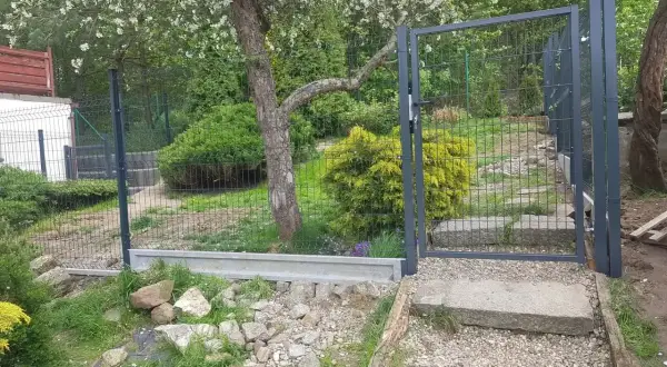 Nowe ogrodzenie z furtką, prawdopodobnie 120 cm wysokości, otacza zielony ogród z kwitnącym drzewem.