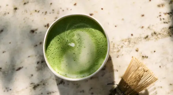 Zielone matcha w filiżance obok bambusowej miotełki na marmurowym blacie.
