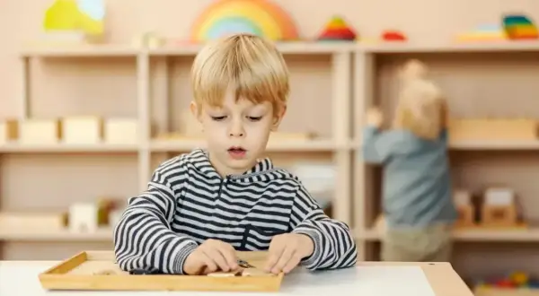Chłopiec skupiony na zadaniu z materiałami Montessori. W tle inne dziecko i regały z zabawkami.
