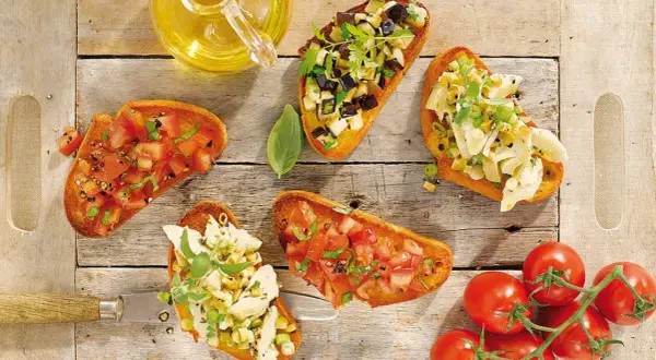 Fünf Bruschetta-Varianten mit Tomaten, Auberginen und Artischocken auf Holzhintergrund.