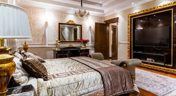 Elegancka sypialnia glamour beżowa z bogato zdobionym łóżkiem, kryształowym żyrandolem i złotymi detalami.