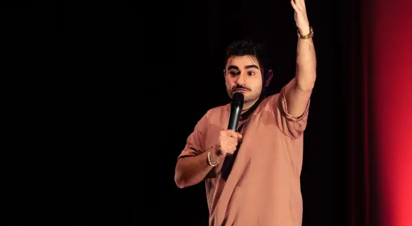 David Puerto Zaragoza, un comediante, levanta la mano mientras sostiene un micrófono en un escenario con cortinas rojas.
