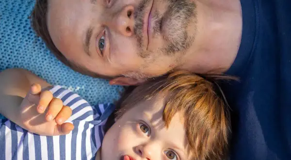 Un padre e il suo bambino con sindrome di Down, un neonato con un'espressione giocosa, sdraiati su una coperta blu.