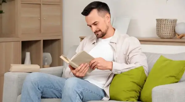 Mężczyzna w jasnej koszuli i jeansach siedzi na kanapie i czyta książkę.
