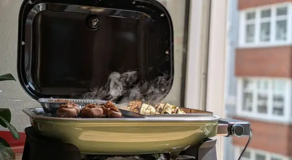 Leckeres Grillen auf dem Balkon: Gefüllte Champignons und Spieße garen in einem grünen Weber Lumin Grill. Rauch steigt auf.