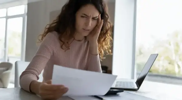 Zmartwiona kobieta analizuje dokumenty, rozważając odrzucenie spadku. Na stole leży laptop i kalkulator.