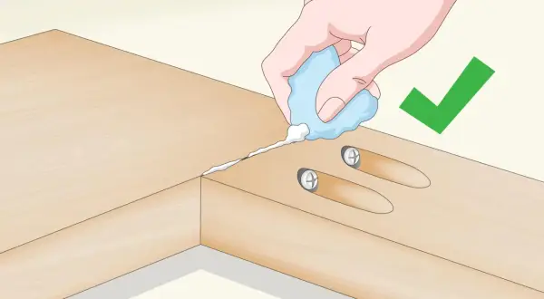 Application de colle pour assembler 2 planches en longueur. Une main tient un tube de colle, deux vis sont déjà en place.