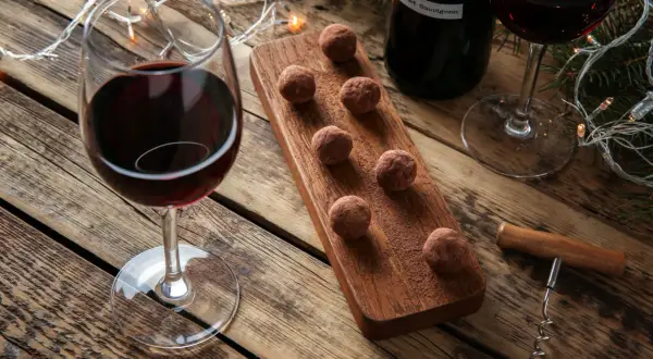 Ein Glas Rotwein und Schokolade-Trüffel auf einem Holzbrett, daneben eine Flasche Rotwein und Lichterketten. Perfekt für gemütliche Abende.