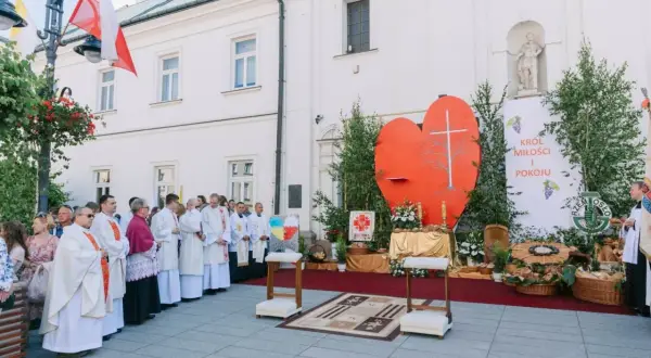 Uroczysta procesja Bożego Ciała przed kościołem. Wierni w białych szatach, wielkie serce z krzyżem i transparent "Król Miłości i Pokoju".