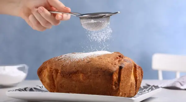 Una mano setaccia zucchero a velo su un dolce appena sfornato, un tocco finale perfetto per ogni ricetta Bimby.