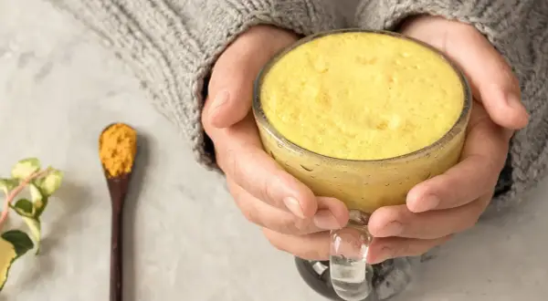 Warme goldene Milch in einer Tasse, gehalten von Händen in einem Strickpullover. Daneben ein Löffel mit Kurkumapulver. Die goldene Milch Wirkung ist bekannt für ihre entzündungshemmenden Eigenschaften.